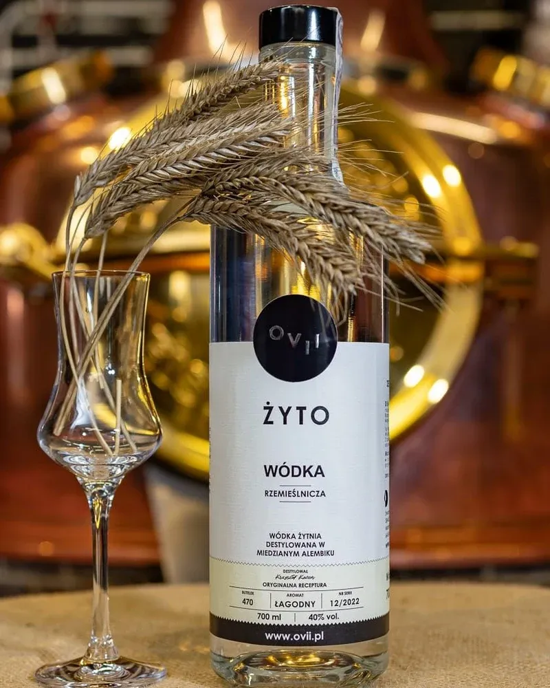 Butelka OVII wódka żytnia z kłosami żyta i kieliszkiem, na tle miedzianego alembiku